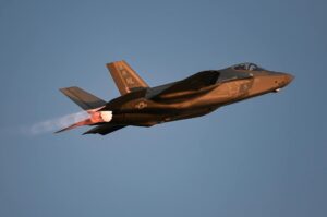 A military jet glides through the air with afterburners glowing at dusk against a clear sky.