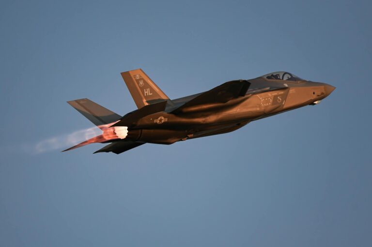A military jet glides through the air with afterburners glowing at dusk against a clear sky.